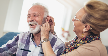 An elderly woman adjusts the hearing aid of an elderly man.
