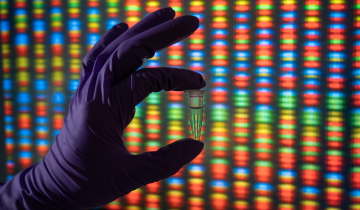 A gloved hand holding a vial in front of a genome sequencing display.