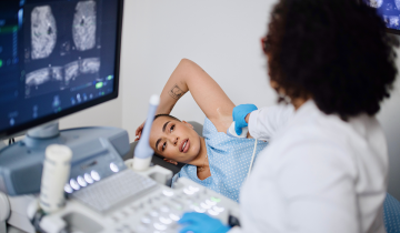 A medical professional conducts a breast cancer screening ultrasound on a woman, with the ultrasound images displayed on a monitor in a clinic setting.