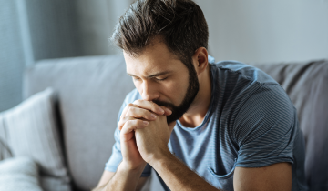 A man sits with his elbows on his knees and hands clasped, looking thoughtful.